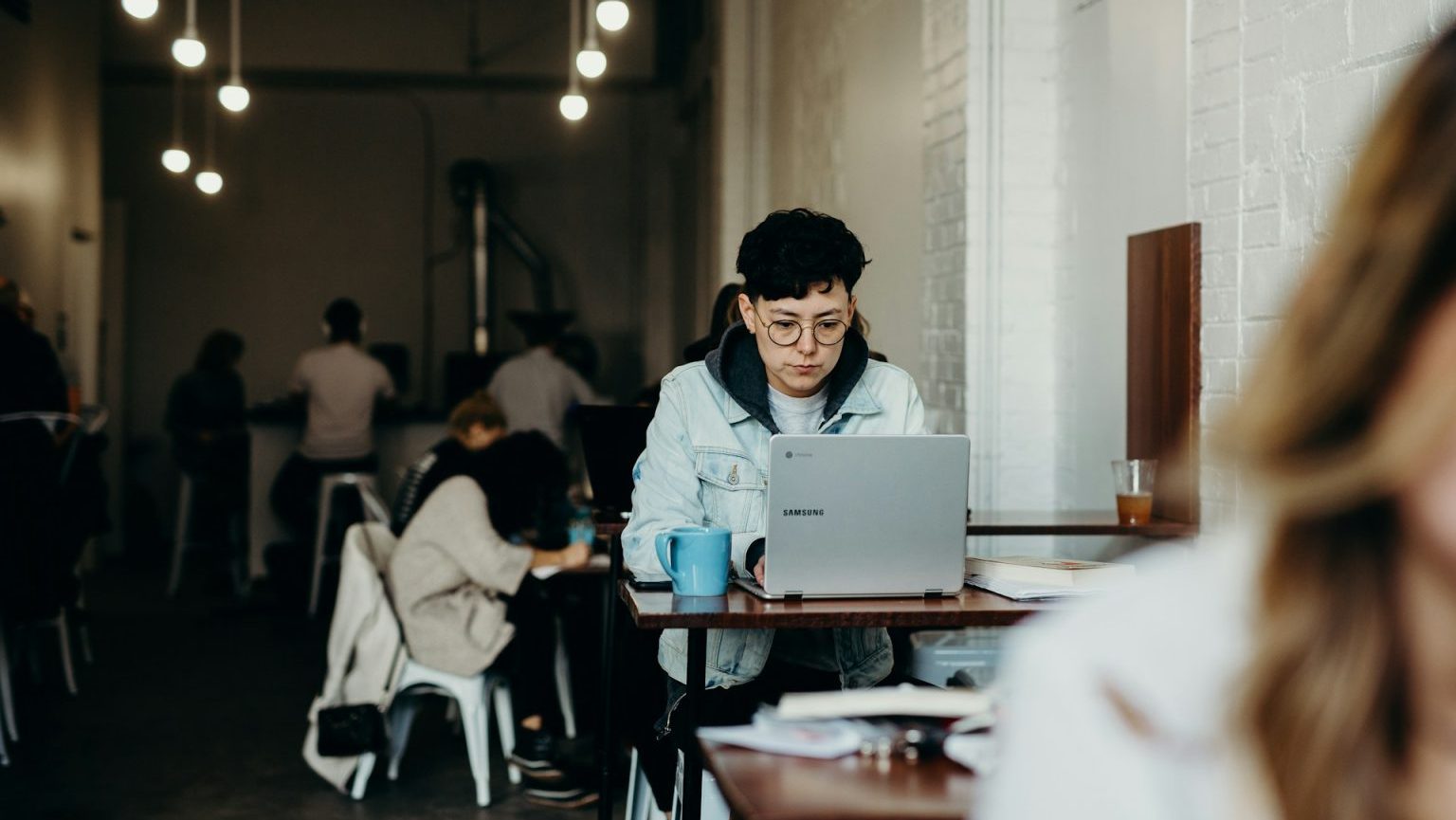 A person wearing glasses and a denim jacket works on a laptop at a table in a cafe. Several other individuals, including both humans and AI enthusiasts, are seated and engaged in different activities in the background.