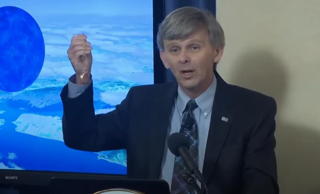 A man in a suit holding a vial while giving a presentation, with a blue globe graphic illustrating gold origin in the background.