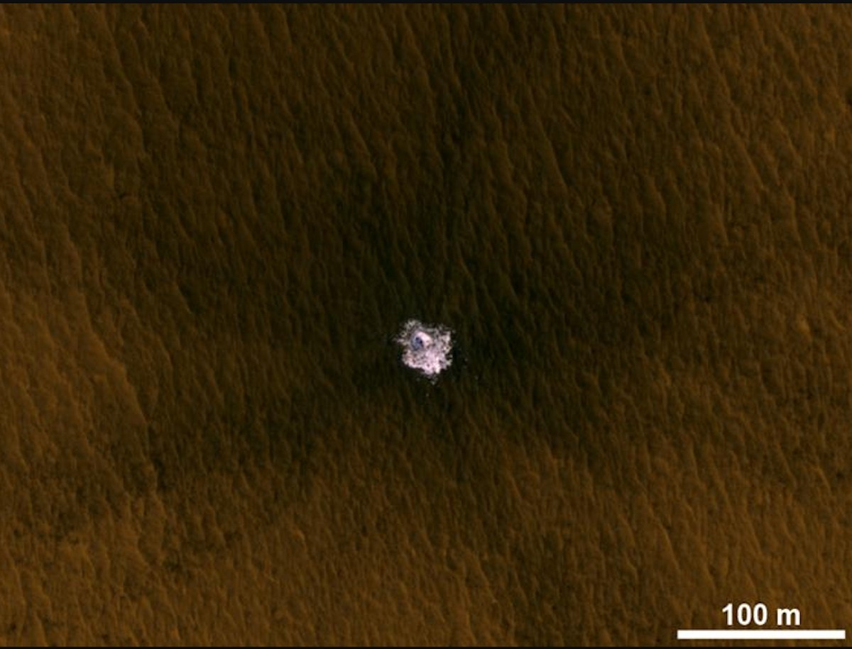 Aerial view of an impact crater on a brown landscape, showcasing a central white area where the impact occurred. A scale bar indicating 100 meters is present in the bottom right corner.