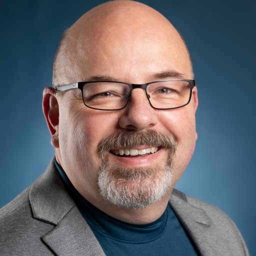 Bald man with glasses, a goatee, and a mustache smiles while wearing a gray blazer and a teal shirt against a blue background.