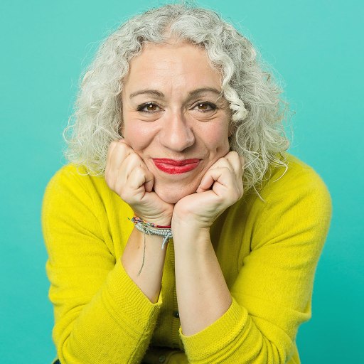 A person with curly gray hair and wearing a yellow sweater rests their head on their hands, smiling against a teal background.