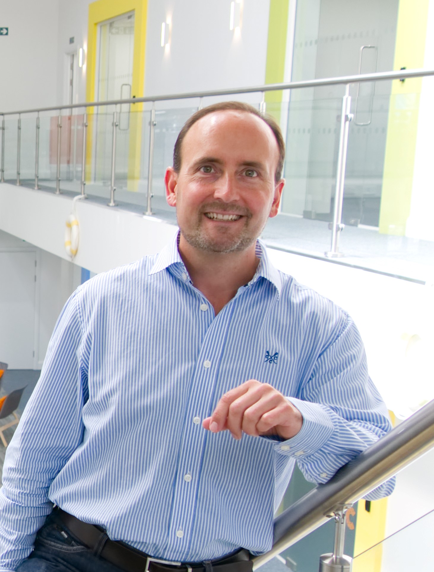 A man in a blue striped shirt and black pants stands leaning on a railing indoors, smiling at the camera.