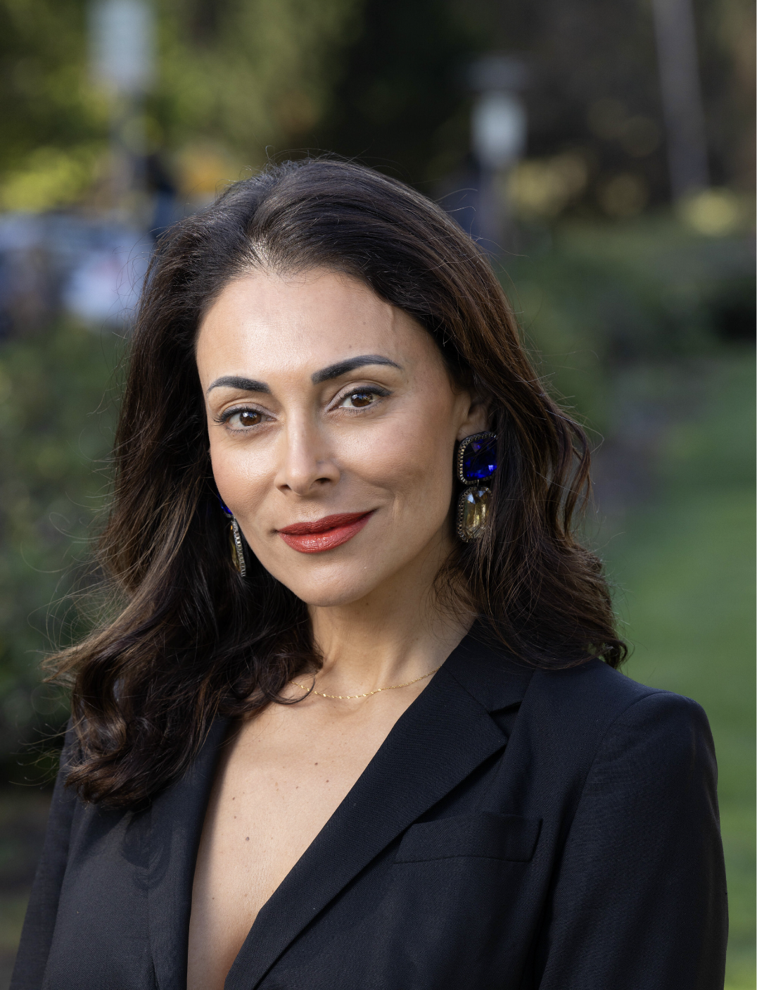 A woman with dark hair wearing a black blazer, large blue earrings, and light makeup, standing outdoors with a blurred green background.