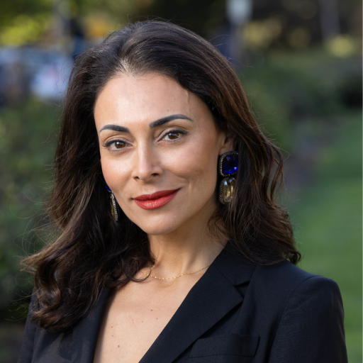 A woman with dark hair wearing a black blazer, large blue earrings, and light makeup, standing outdoors with a blurred green background.