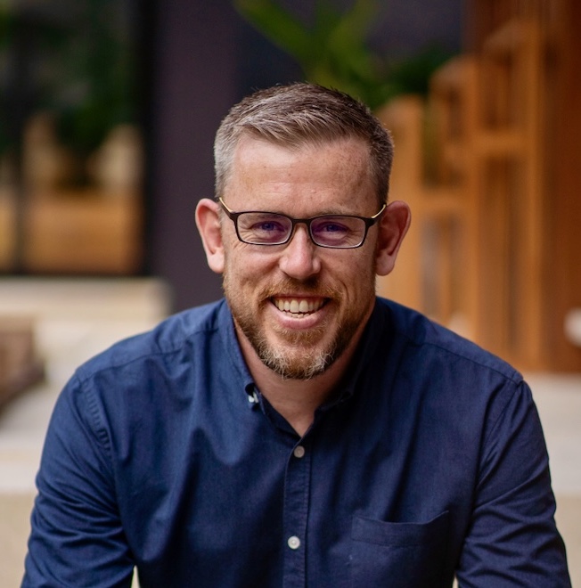 A man with short, light brown hair and glasses, wearing a navy blue button-up shirt, is smiling and looking at the camera.