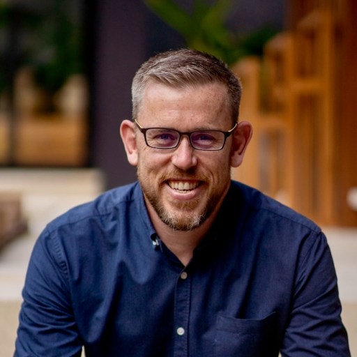 A man with short, light brown hair and glasses, wearing a navy blue button-up shirt, is smiling and looking at the camera.