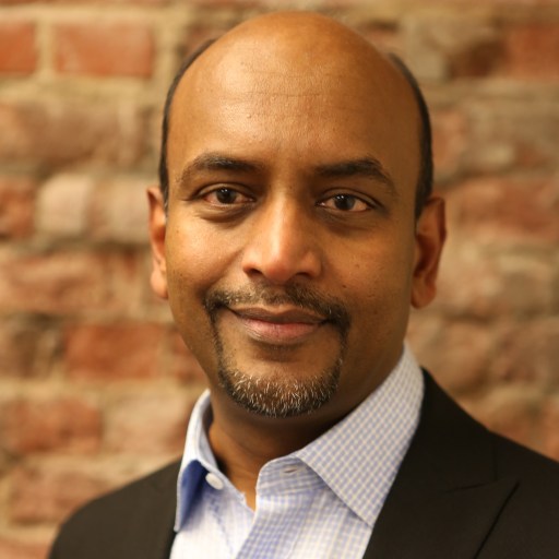 A man with a shaved head, wearing a suit jacket and blue shirt, stands in front of a brick wall, smiling slightly.