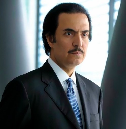 A man in a suit and tie stands indoors against a backdrop of vertical blinds.