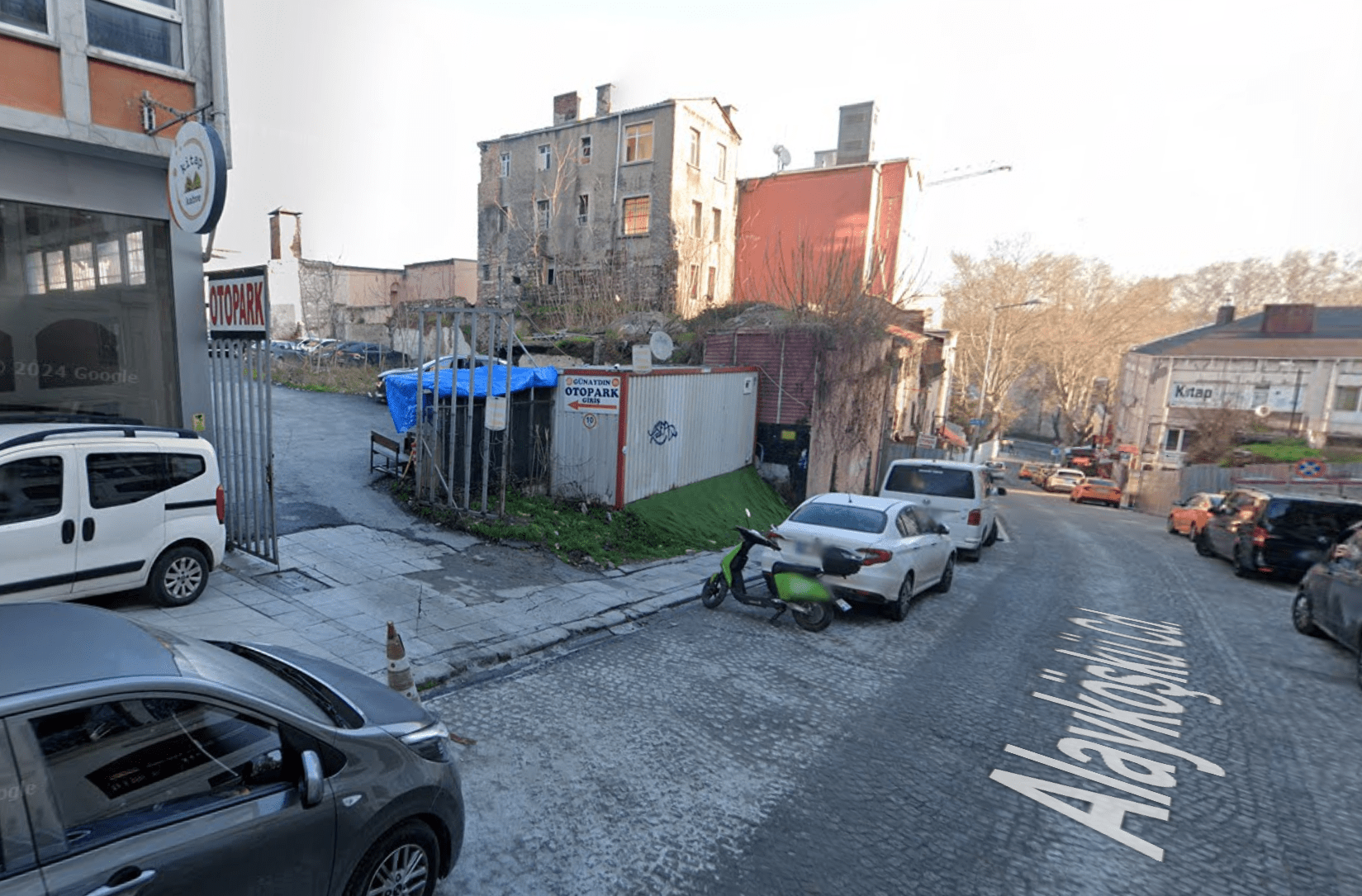 A cobblestone street with parked vehicles, a small gated lot with a tent, and surrounding buildings, taken near the junction of Alayköşkü Cd.