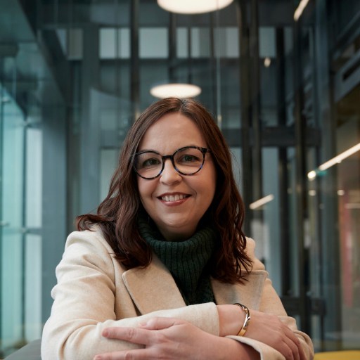 A woman with long brown hair and glasses, wearing a light coat and green turtleneck, smiles while seated indoors with modern, glass-walled surroundings.