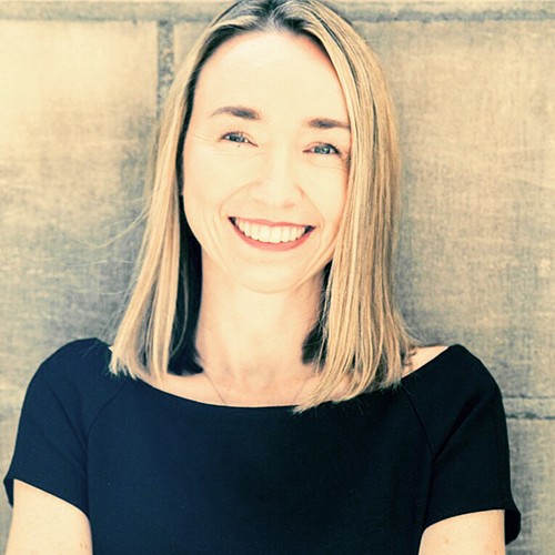 A woman with straight, shoulder-length blonde hair is smiling and wearing a black top against a plain background.