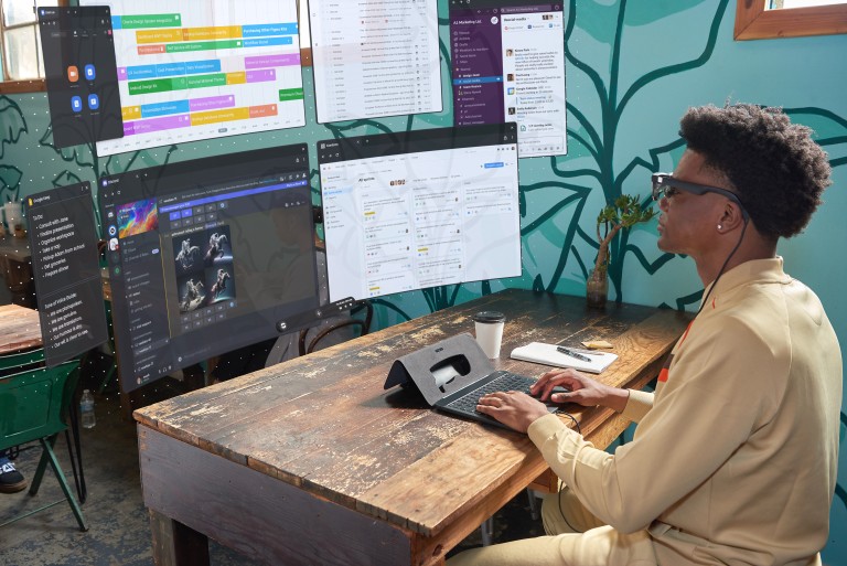 A person sits at a wooden desk using a laptop with multiple virtual screens displaying various apps and documents. A cup of coffee and a phone are also on the desk. The background features a mural.