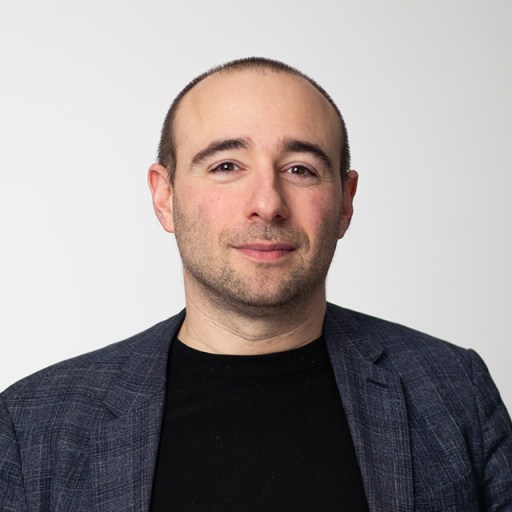 A man with short hair wearing a dark blazer and black shirt stands against a plain white background.