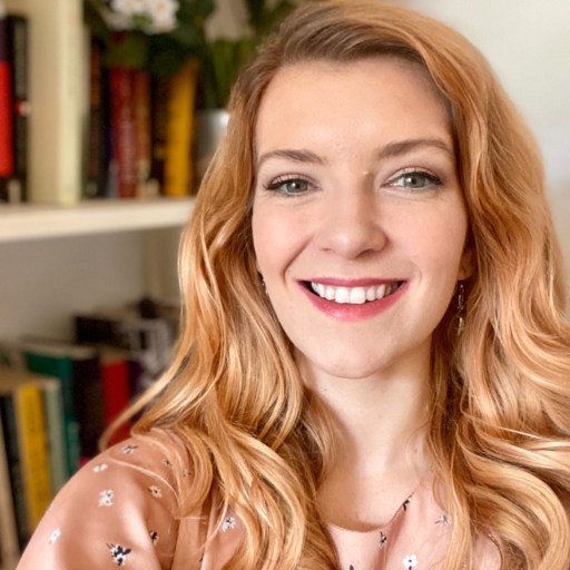 A woman with long wavy hair smiles while posing in front of a bookshelf with colorful books and a vase of flowers.