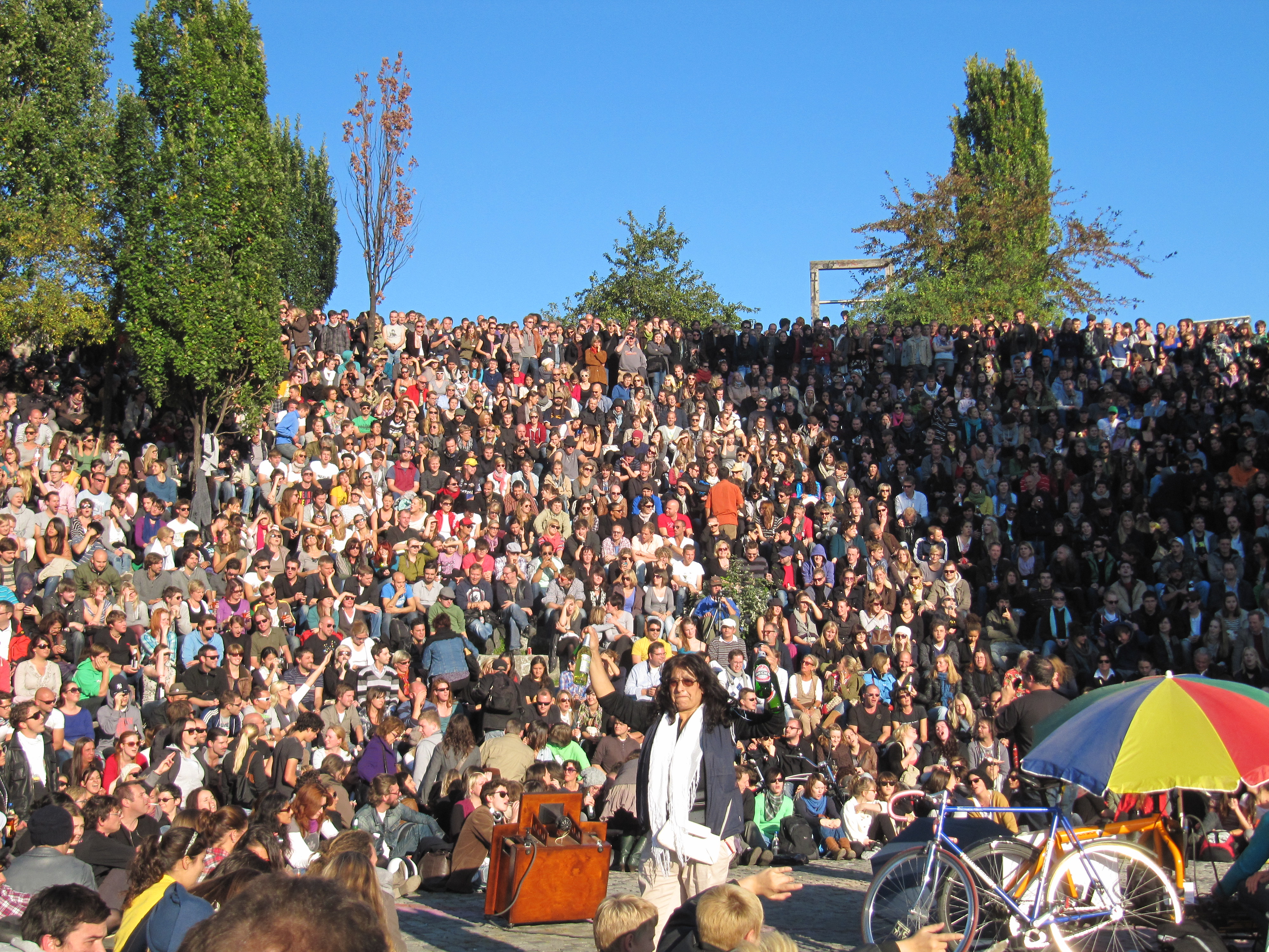 A performer with a suitcase stands before a large, seated crowd in an outdoor amphitheater on a sunny day. An umbrella and a bicycle are visible in the foreground, sparking curiosity about why machines learn from such everyday scenes.