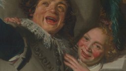 A close-up of two people with rosy cheeks, smiling and laughing together, wearing historical clothing and ornate collars.