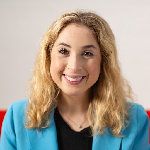 A woman with blonde hair, wearing a blue blazer and a black top, smiles at the camera against a blurred background.