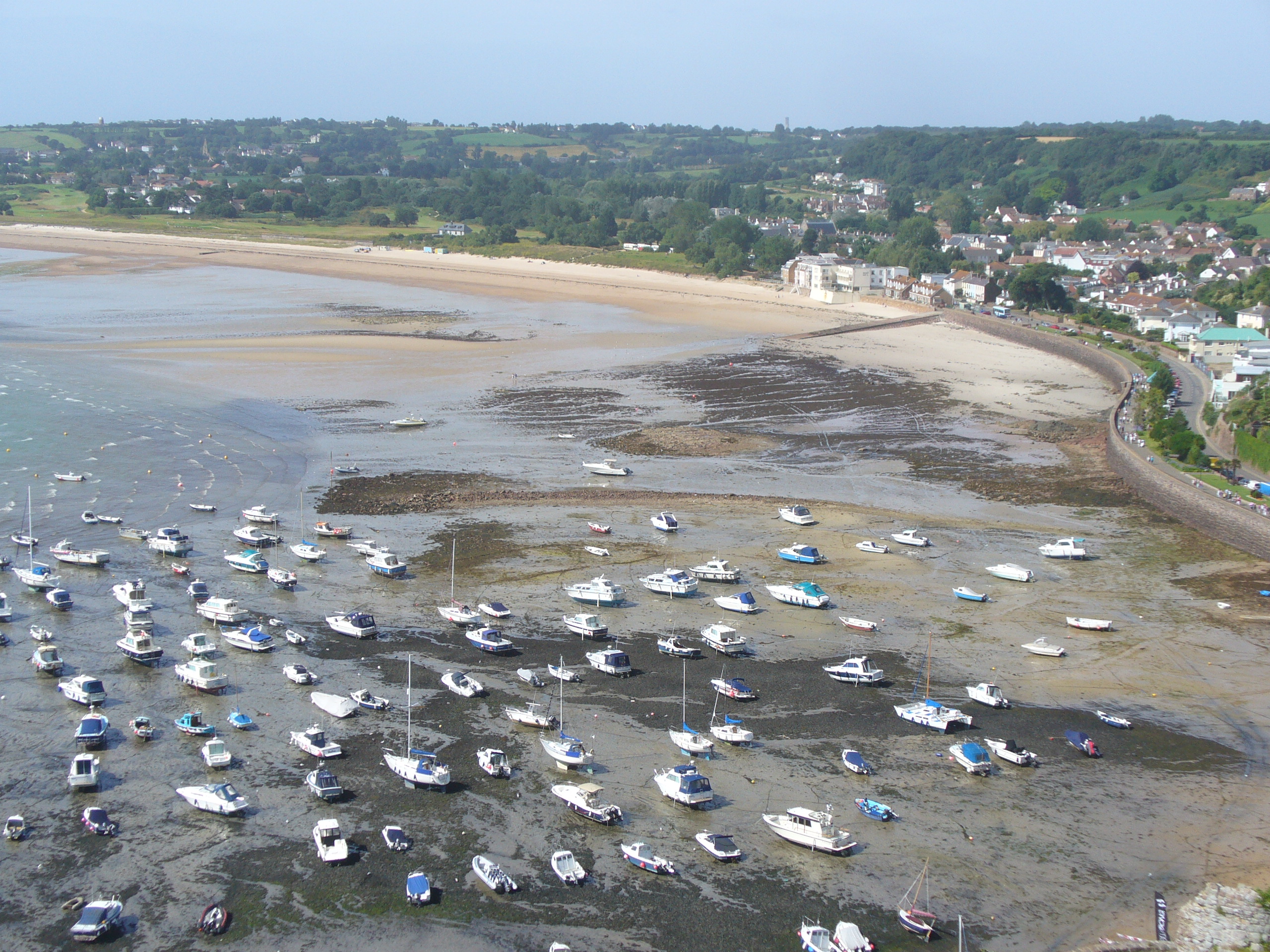 Aerial view of a coastal town with numerous small boats anchored on the water and shore. Houses and greenery are visible in the background, surrounding a sandy beach area.