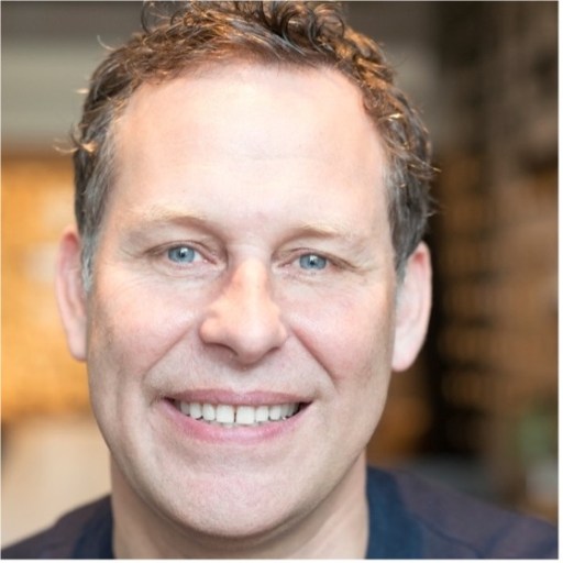 A man with short, curly hair and blue eyes is smiling, looking directly at the camera. He is wearing a dark shirt, and the background is out of focus.