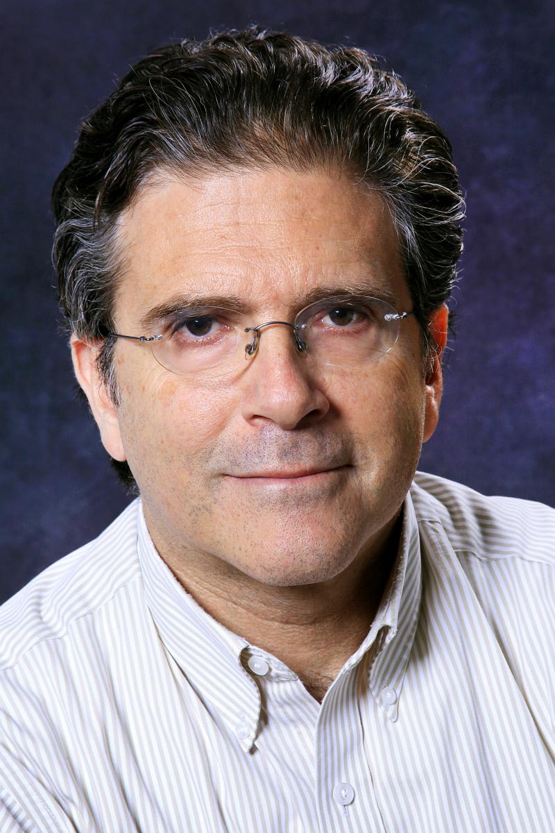Middle-aged man with glasses, dark wavy hair, and a striped button-down shirt, posing against a dark, textured background.