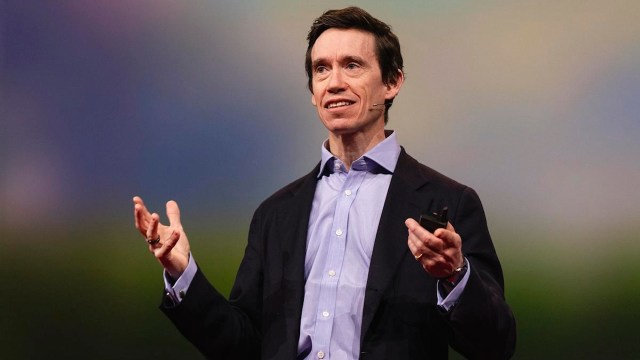 A person in a suit stands on stage, gesturing with both hands while speaking into a headset microphone, passionately discussing direct giving in their presentation.