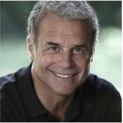 A man with short gray hair and a broad smile is wearing a dark polo shirt, looking directly at the camera against an out-of-focus green background.