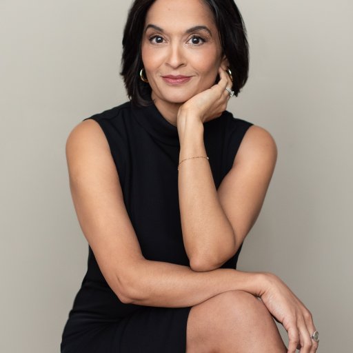 A woman with short black hair is sitting, wearing a black sleeveless dress. She has her chin resting on her hand and she is smiling slightly, with a neutral background.