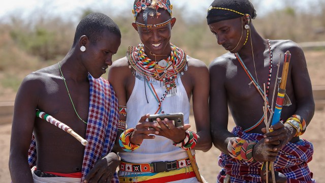 Three Masai men, dressed in traditional attire and adorned with beaded jewelry, gather around a smartphone in an outdoor setting.