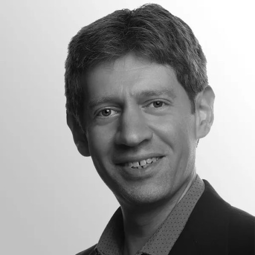 A black and white headshot of a middle-aged person with short hair, a slight smile, wearing a collared shirt and a dark blazer.