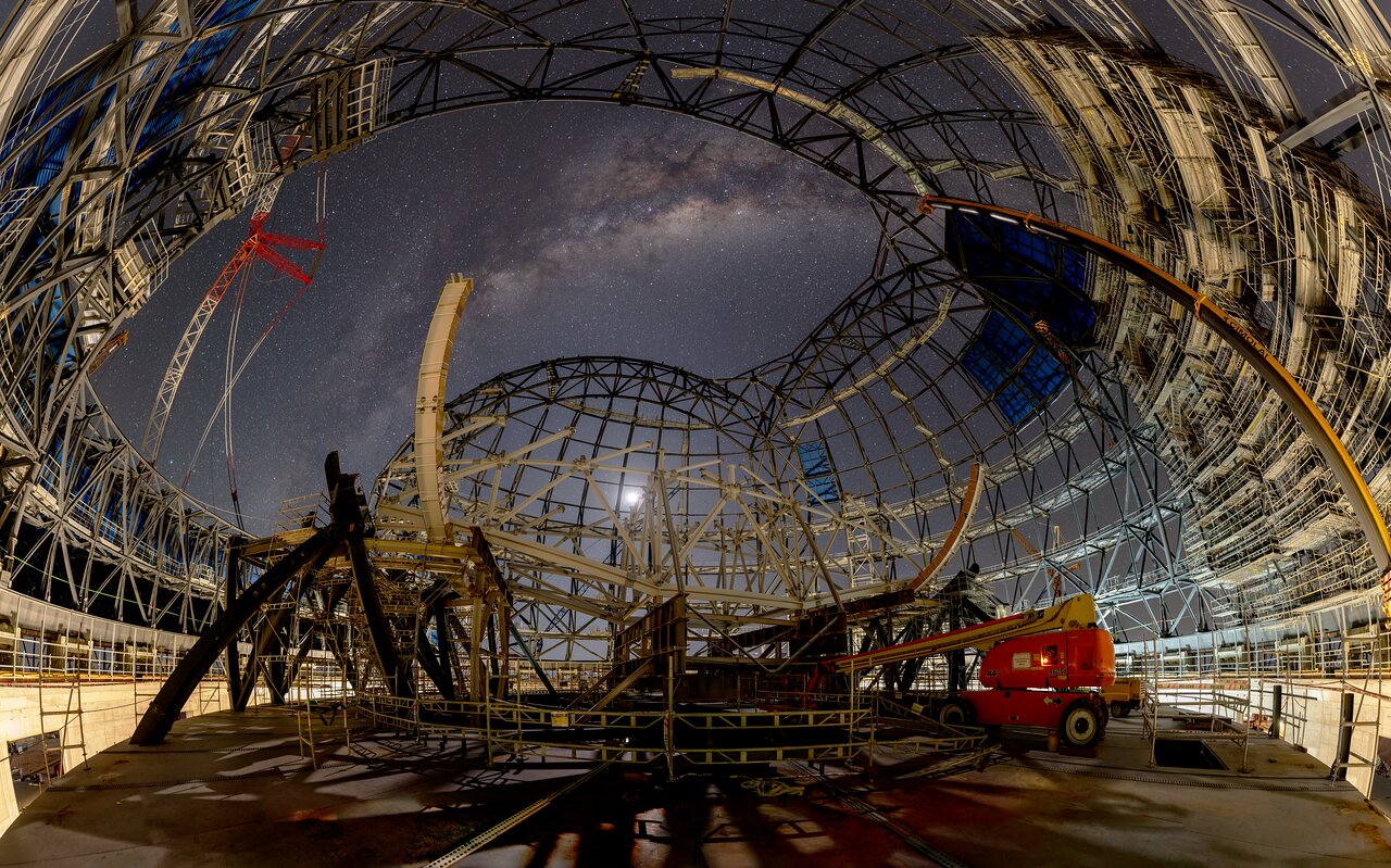 A large, open-structure telescope under construction at night with the Milky Way visible in the sky above and a red crane on the left side.