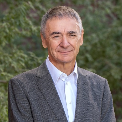 A man with short gray hair wearing a gray suit and white shirt stands outdoors, with green foliage in the background.