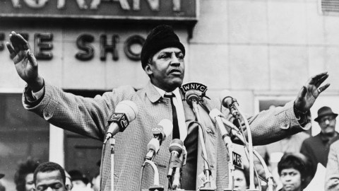 A man standing behind multiple microphones gestures while speaking to a crowd in an outdoor setting, with a group of people and a building in the background.