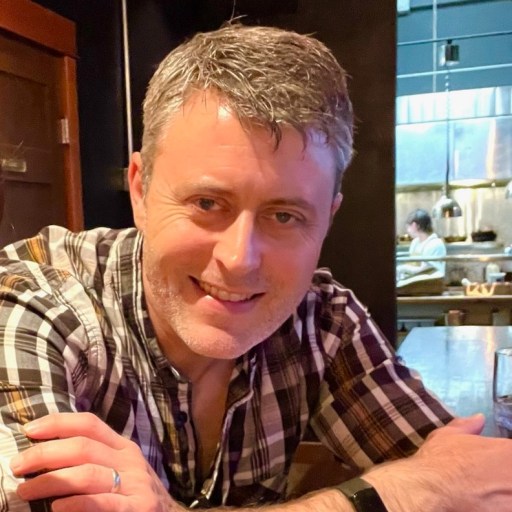 A man with short gray hair and wearing a plaid shirt is seated at a wooden table in a dimly lit restaurant, smiling at the camera.