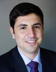 A man with dark hair and a smile, wearing a dark suit, light blue shirt, and purple tie, is pictured against a neutral background.