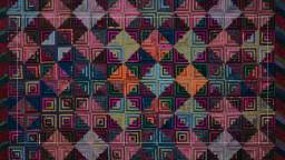 A colorful geometric quilt features a complex pattern of interlocking squares and rectangles in shades of pink, yellow, blue, and green.