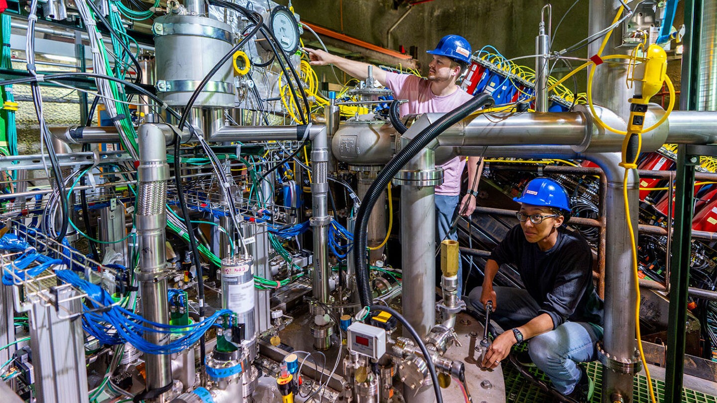 Two individuals in hard hats and safety glasses working on complex machinery with numerous cables and metal components in an industrial setting.