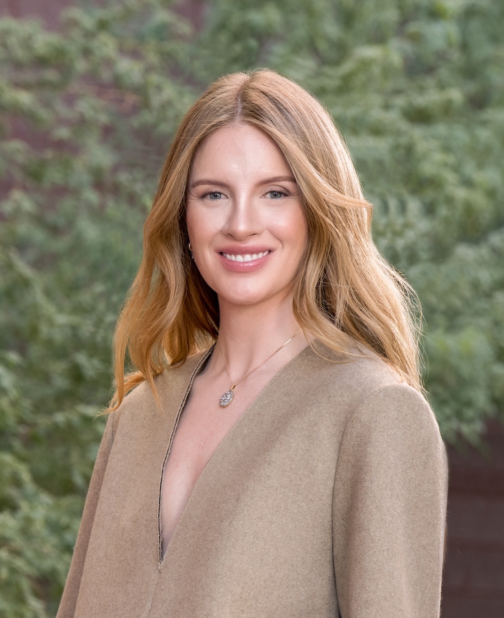 A woman with long, light brown hair wearing a beige coat poses outdoors in front of greenery.