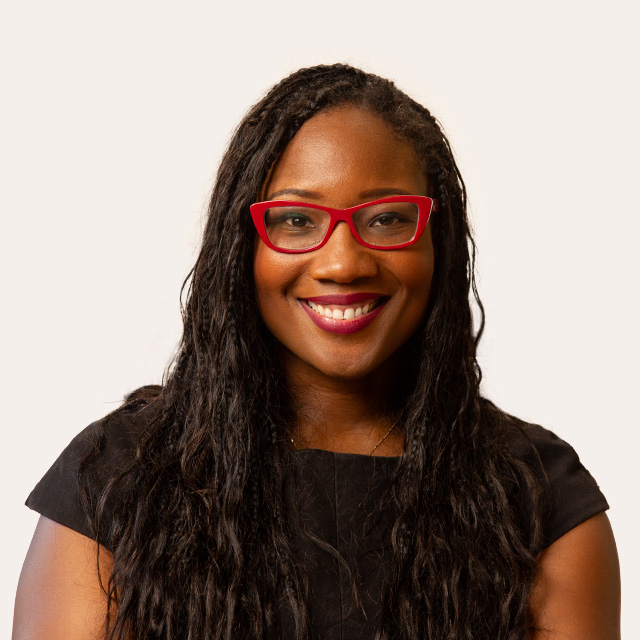 A person with long dark wavy hair, wearing red glasses and a black top, smiles against a plain background.