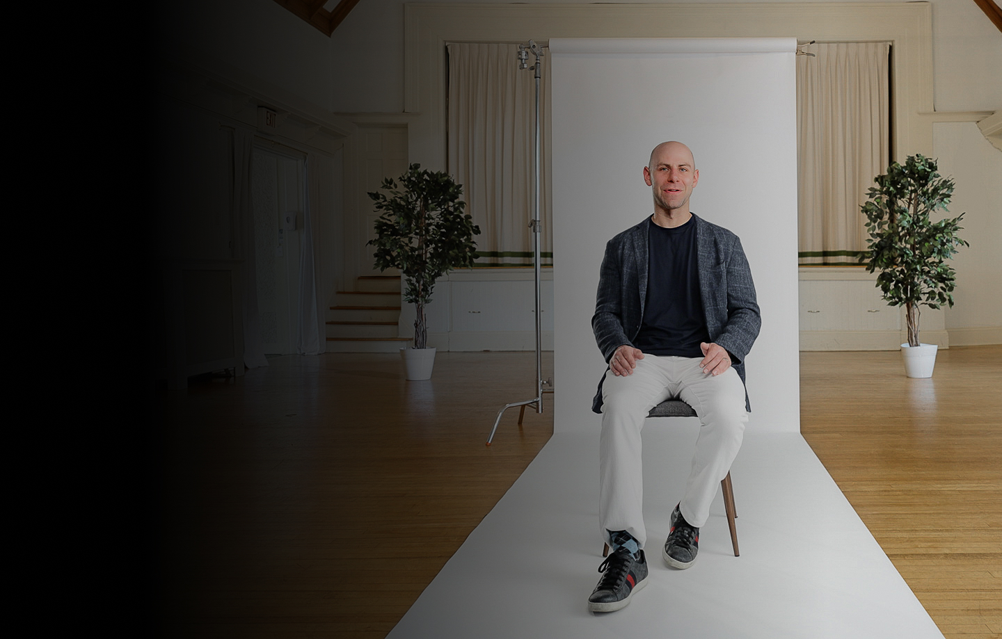 A man sits on a chair in front of a white backdrop, flanked by two potted plants, in a well-lit room with a wooden floor.