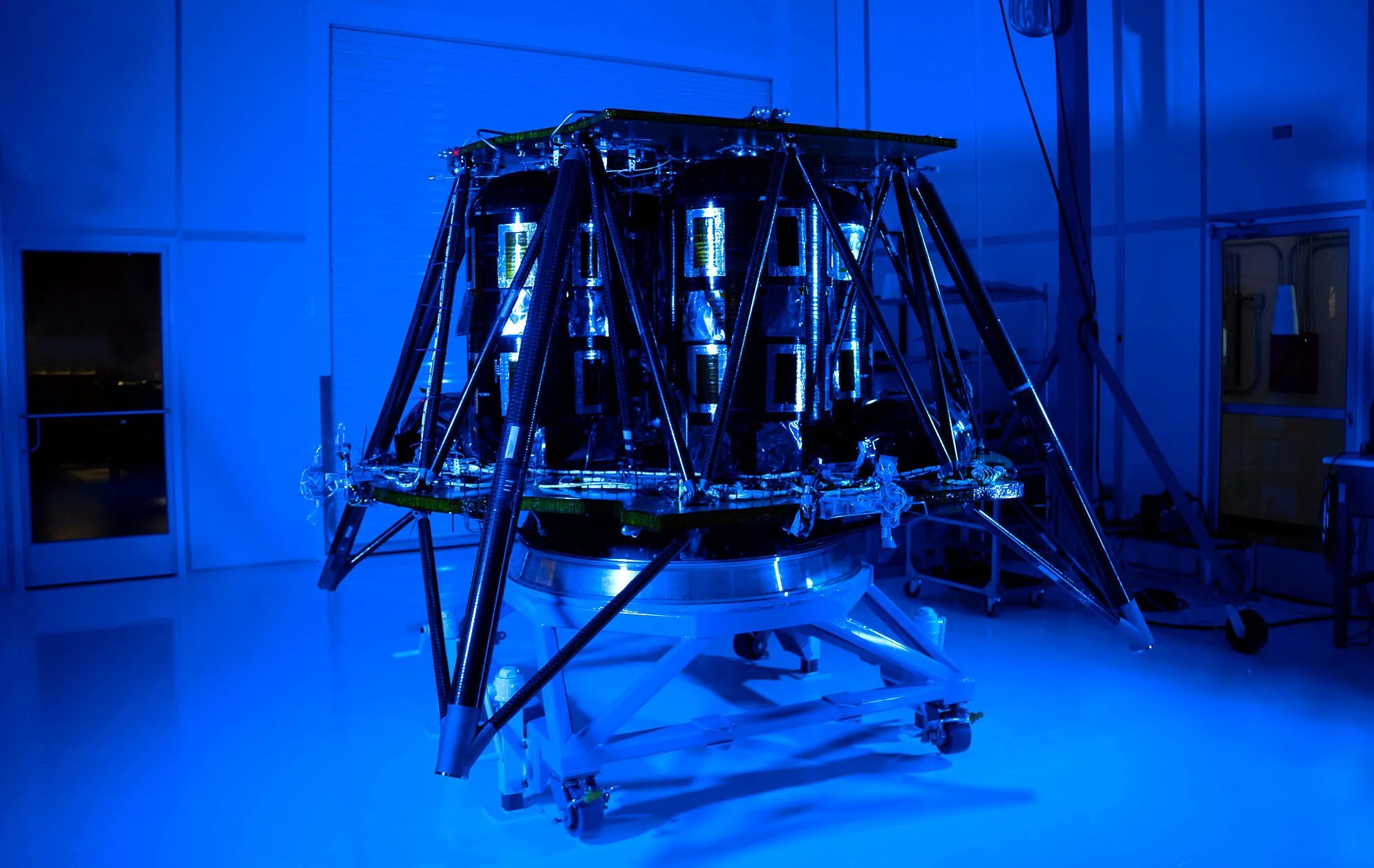 A spacecraft assembly in a dimly lit, blue-illuminated cleanroom, featuring intricate structures and supports, mounted on a wheeled platform.