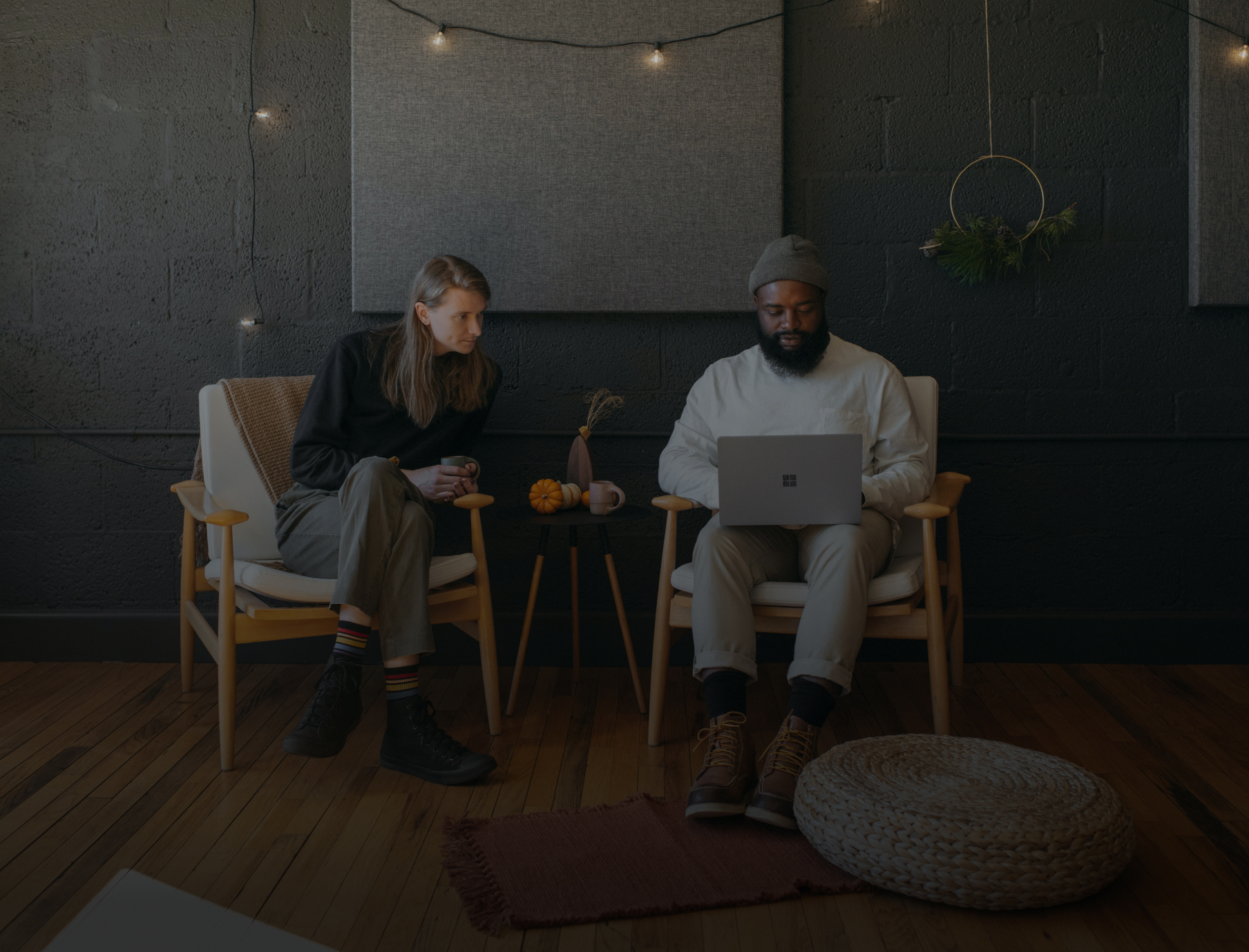 Two people sitting in chairs with a laptop in a cozy room, featuring string lights and wall decor.