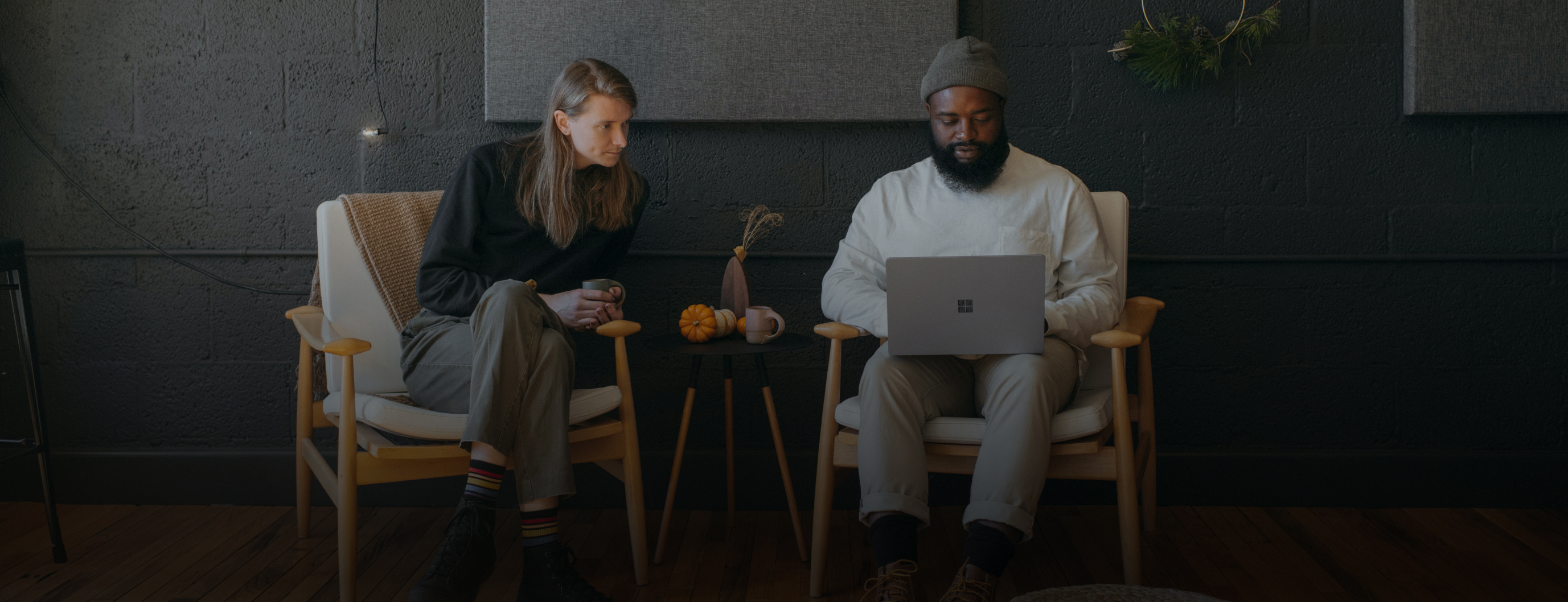 Two people sitting on chairs, one holding a cup and the other using a laptop. A small table with a vase and cups is between them. The background is a dark wall with acoustic panels.