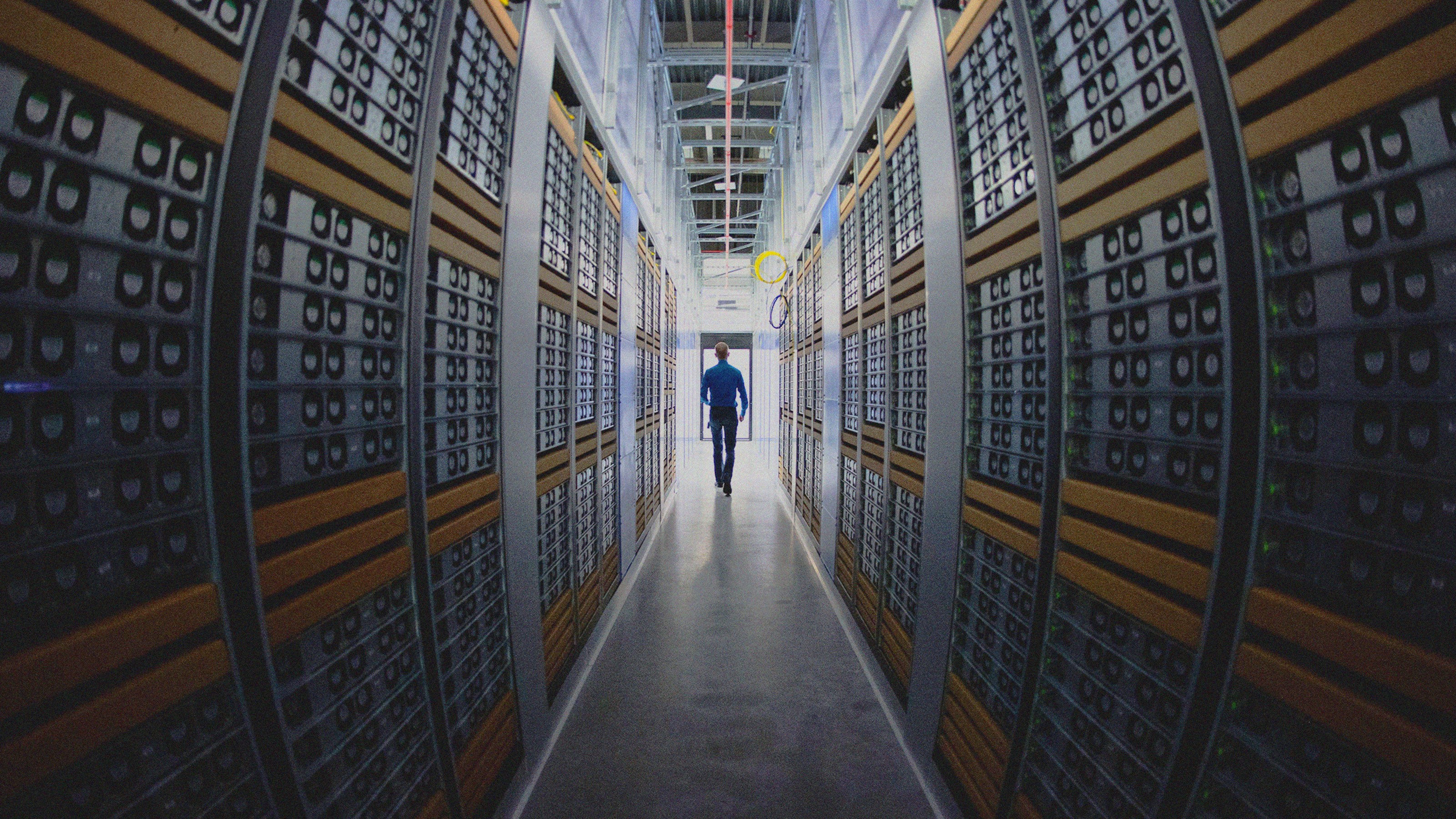 A wide shot of a man walking down an aisle between tall stacks of server racks in a data center filled with computer hardware.