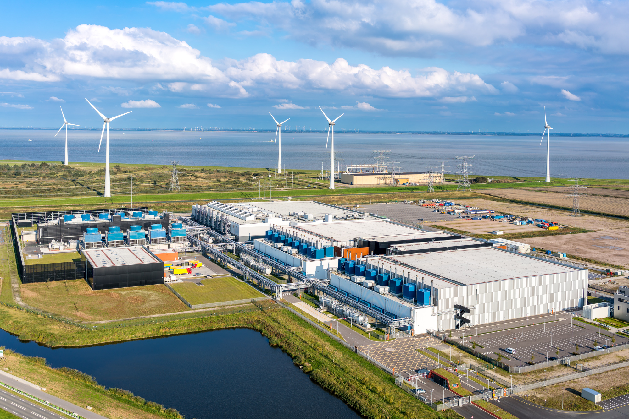 Aerial view of a coastal industrial complex with large buildings, wind turbines, and a parking area near a body of water under partly cloudy skies.