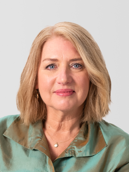 A woman with shoulder-length blonde hair is wearing a green blouse and a small necklace with a pendant, posing against a plain background.