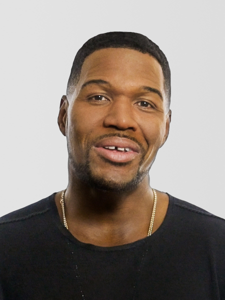 A person with short hair and a neatly trimmed beard smiles while wearing a black shirt and a gold chain necklace against a plain light-gray background.