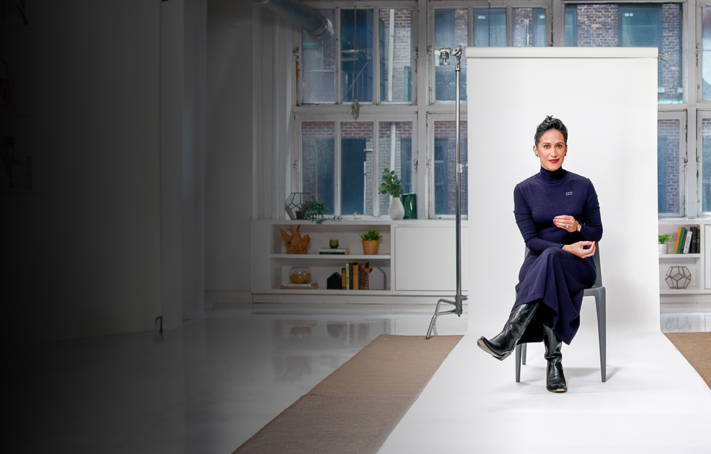 A woman in a dark dress and boots sits on a chair in a well-lit studio with a white backdrop and large windows in the background.