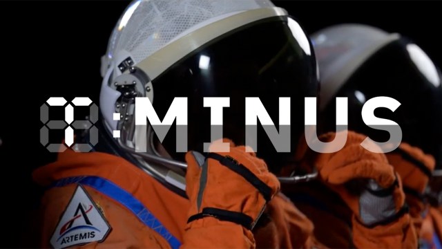 Two astronauts in orange spacesuits and helmets prepare for a mission. A digital countdown displays "T-MINUS," with an Artemis mission patch visible on one spacesuit.