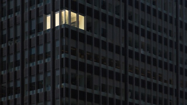 A high-rise office building at night with one illuminated corner office visible amidst the darkened windows, a lone workaholic burning the midnight oil.