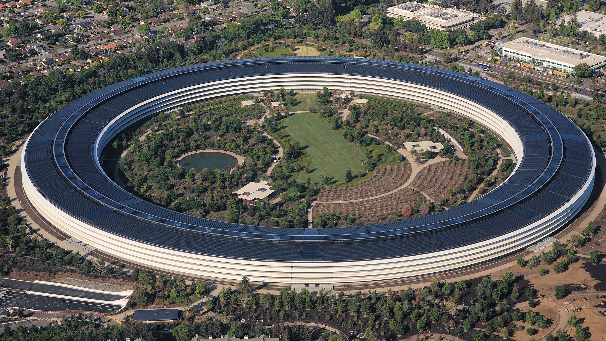 Aerial view of a circular office building surrounded by trees, with a landscaped garden and pond in the center, located in a suburban area with residential homes and other buildings nearby.